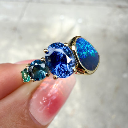 An Australian Opal Ring by NIXIN Jewelry. A close-up view of the ring shows the beautiful contrast between the black opal and the saturated blue and teal sapphires and emerald in a modern opal ring design.