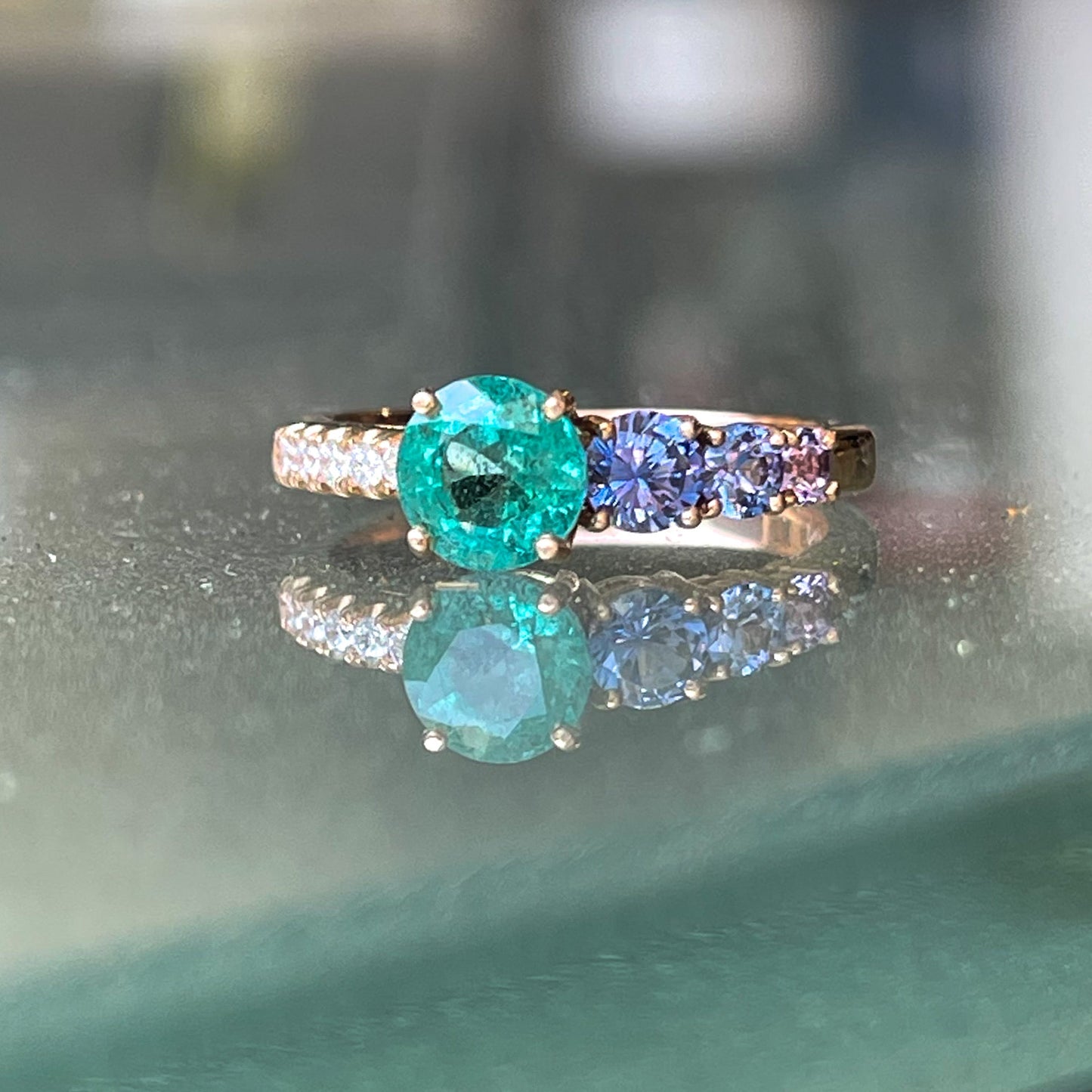 A Colombian Emerald Ring by NIXIN Jewelry rests upon a glass table. The modern emerald ring design features pave diamonds and purple sapphires flanking the green emerald.