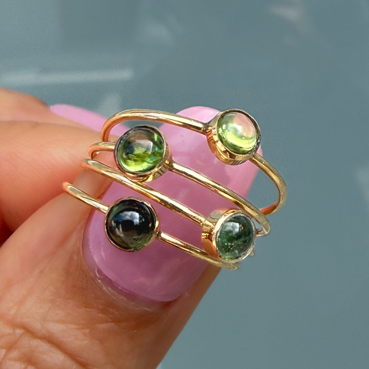 Lucky Green Tourmaline Rings held on a fingertip, four 14k gold stacking rings showing the range of green tourmaline hues.