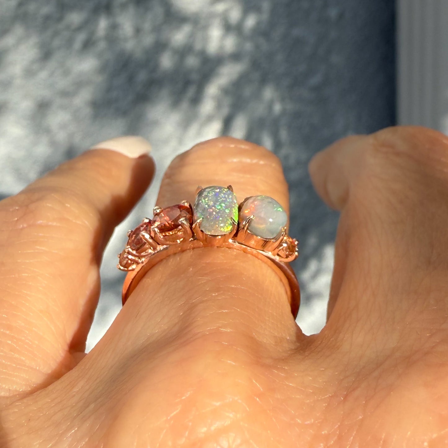 An Australian Opal Ring by NIXIN Jewelry worn on the finger, showing the full scale of this five-stone women's luxury ring cluster. The contrast between the semi black opals and the peach sapphires makes this a standout unique jewelry design. This gorgeous rose gold opal ring is a luxury purchase.