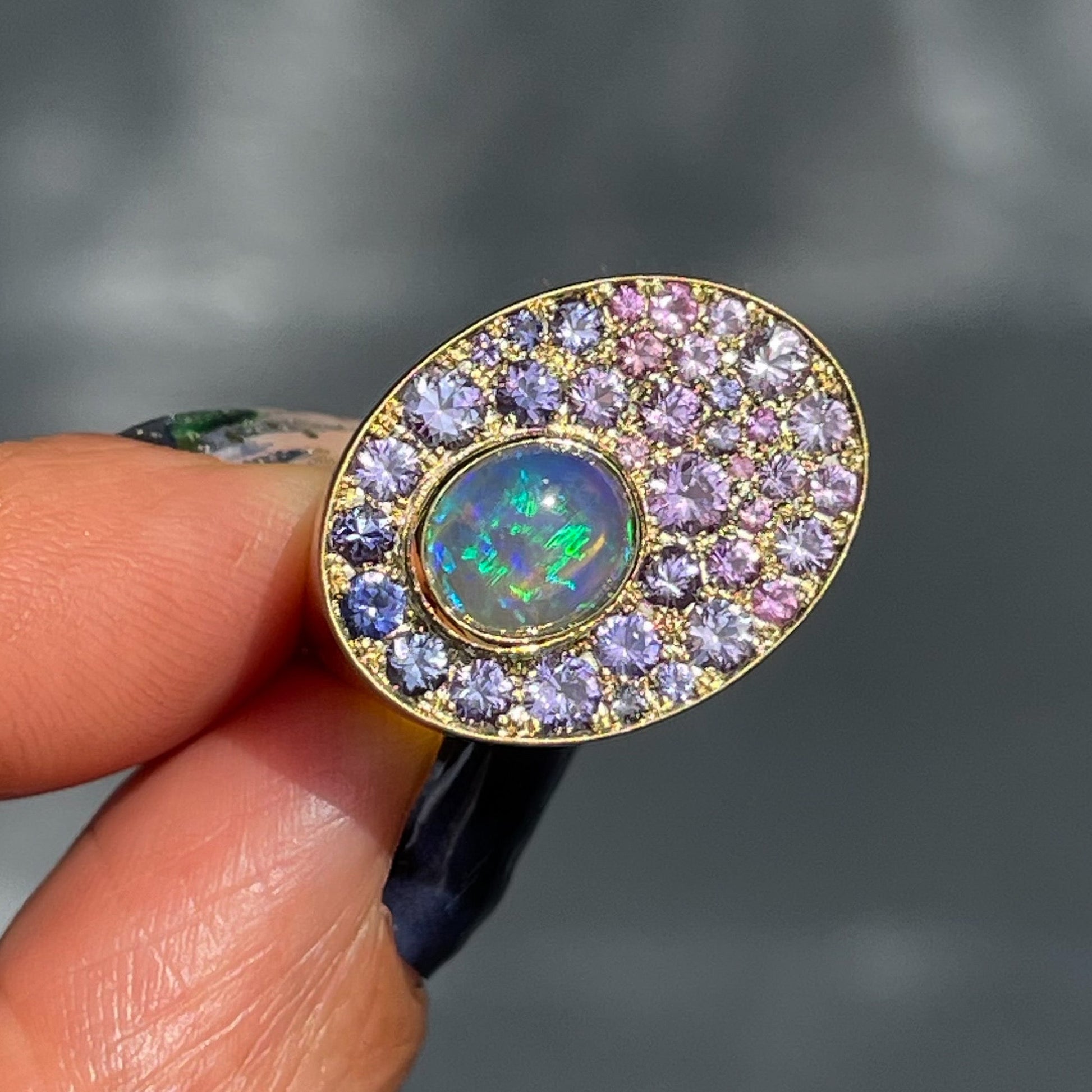 An Australian Opal Ring by NIXIN Jewelry held in front of a grey wall. The sapphire and opal ring is 14k gold.