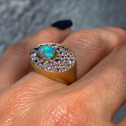 An Australian Opal Ring by NIXIN Jewelry is shown at an angle worn on the hand. The green opal flashes against the gold of the signet ring and the pink and purple sapphires.