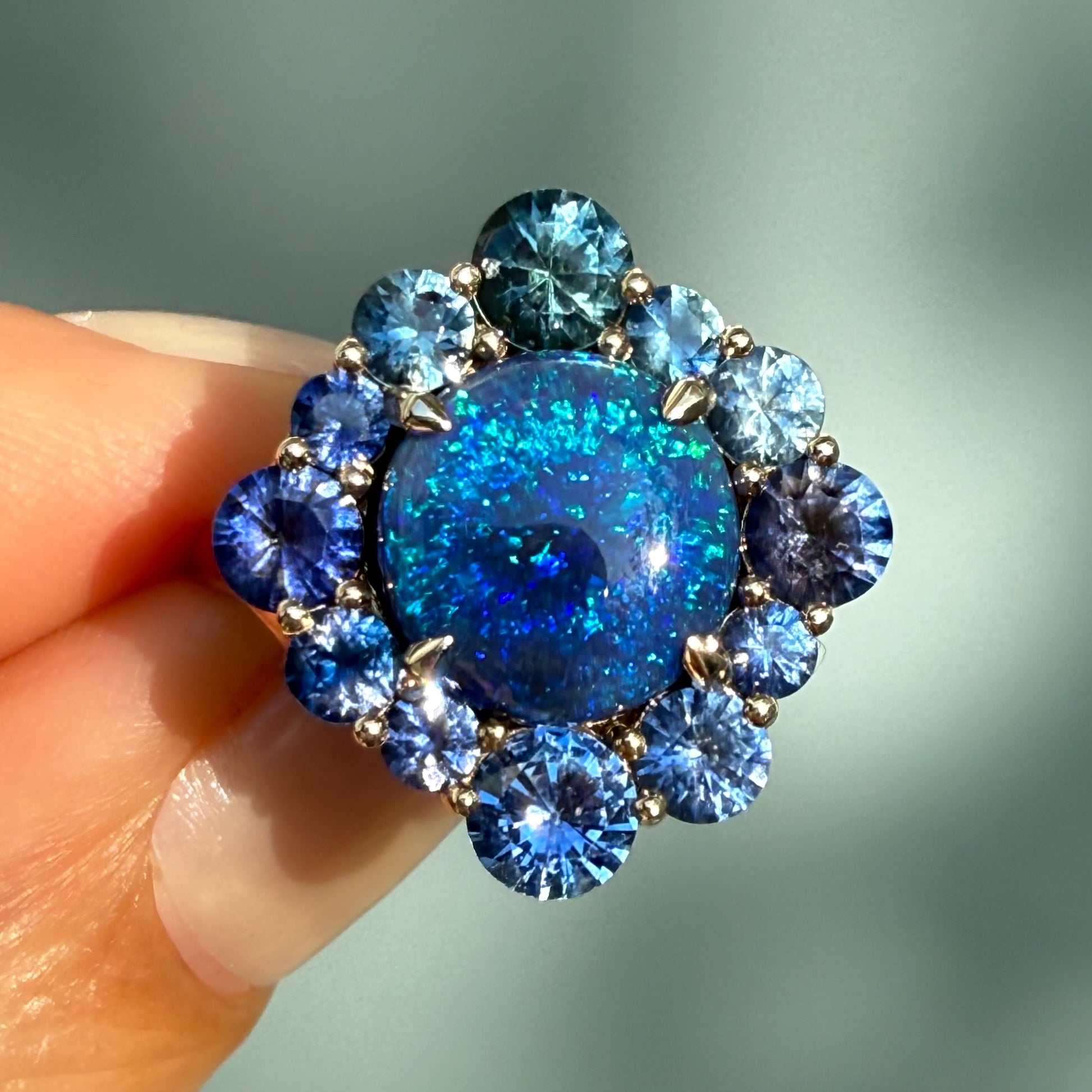 An Australian Opal Ring by NIXIN Jewelry. This unique opal jewelry piece features a round black opal in a prong setting, surrounded by a dynamic halo of sapphires. It is a stunning example of jewelry art and luxury jewelry, perfect for those seeking rare jewelry.