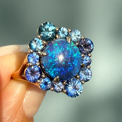 An Australian Opal Ring by NIXIN Jewelry. This angled shot captures the maximum blue and teal fire of the black opal in its gold prong setting beneath strong light. This is a highly desired piece of unique jewelry combining a natural opal with an artistic sapphire halo, making it a standout modern opal ring and exceptional example of black opal jewelry.
