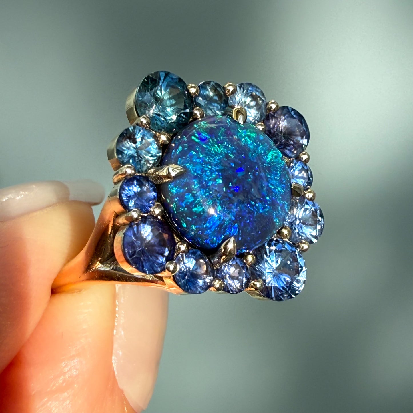 An Australian Opal Ring by NIXIN Jewelry. This black opal jewelry piece is set with a deep australian opal surrounded by an ombre halo of blue and teal sapphires, creating a stunning sapphire opal ring. This impressive piece is considered rare opal jewelry, appealing to collectors searching for a women's luxury ring.