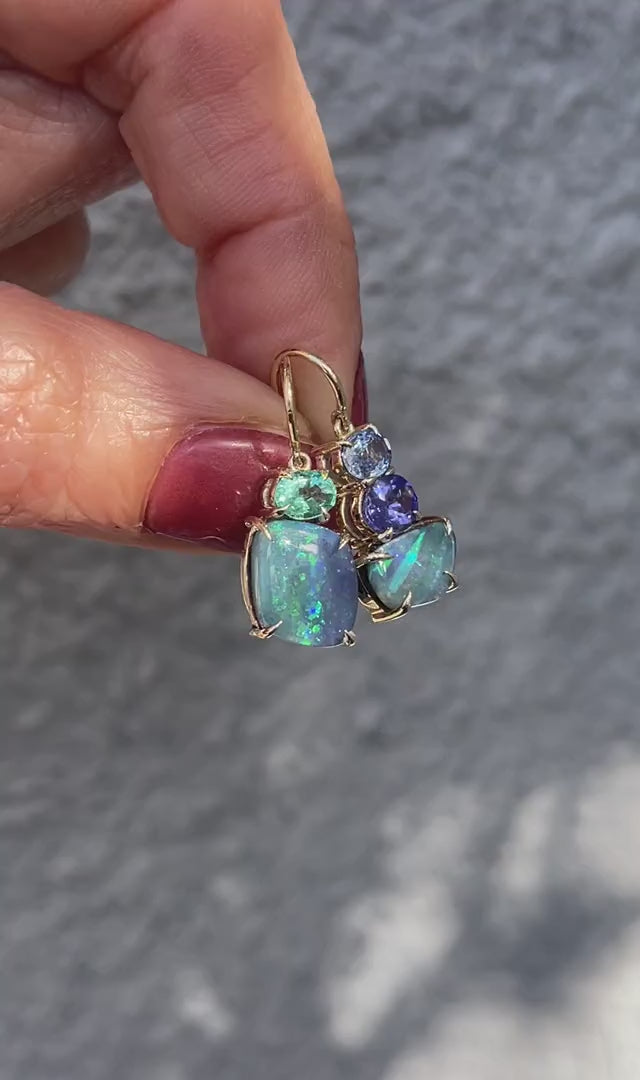 Video of Australian Opal Earrings by NIXIN Jewelry held in front of a grey wall. The opal drop earrings have opals, emerald, sapphire and tanxanite.