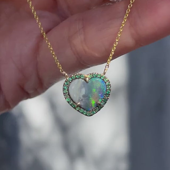 Video of an Australian Opal Heart Necklace by NIXIN Jewelry held in sunlight. The necklace has a heart shaped black opal and emeralds.