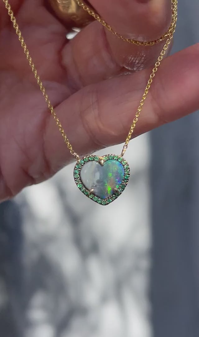 Video of an Australian Opal Heart Necklace by NIXIN Jewelry held in sunlight. The necklace has a heart shaped black opal and emeralds.