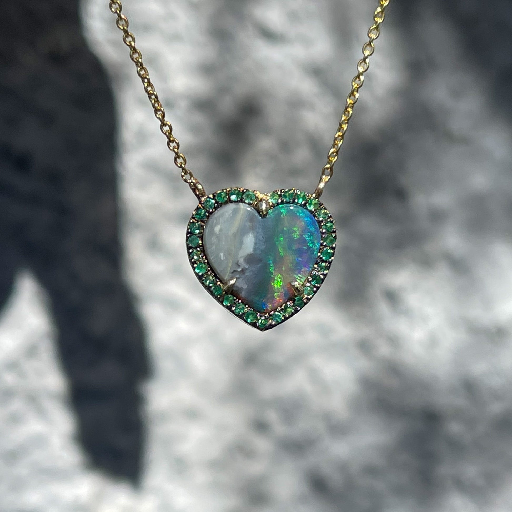 An Australian Opal Heart Necklace by NIXIN Jewelry hanging in front of a grey backdrop. The Black Opal is surrounded by an emerald halo.
