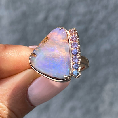 An Australian Opal Ring by NIXIN Jewelry is held in front of a grey wall. The ombre sapphire ring has pink, purple and blue.