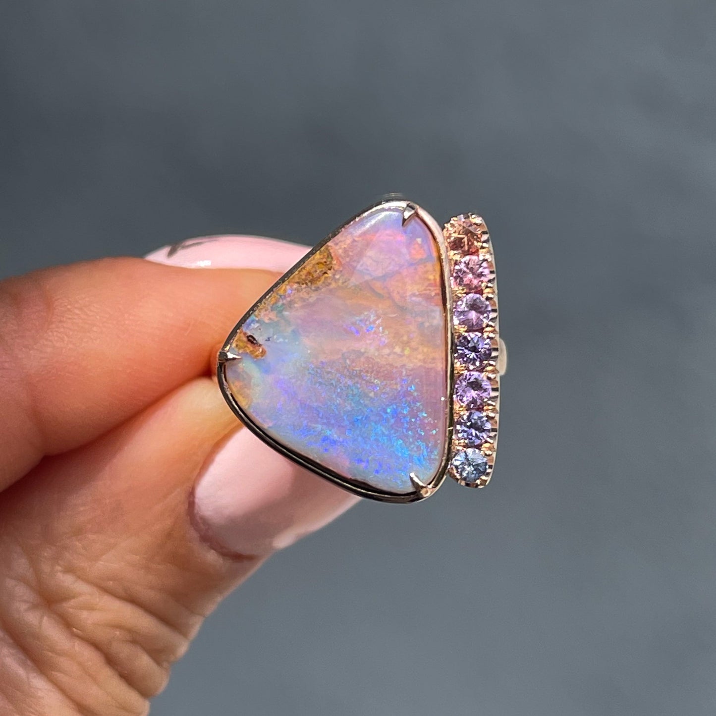 An Australian Opal Ring by NIXIN Jewelry held in front of a grey backdrop. The ring is made with a Boulder Opal and ombré sapphires.