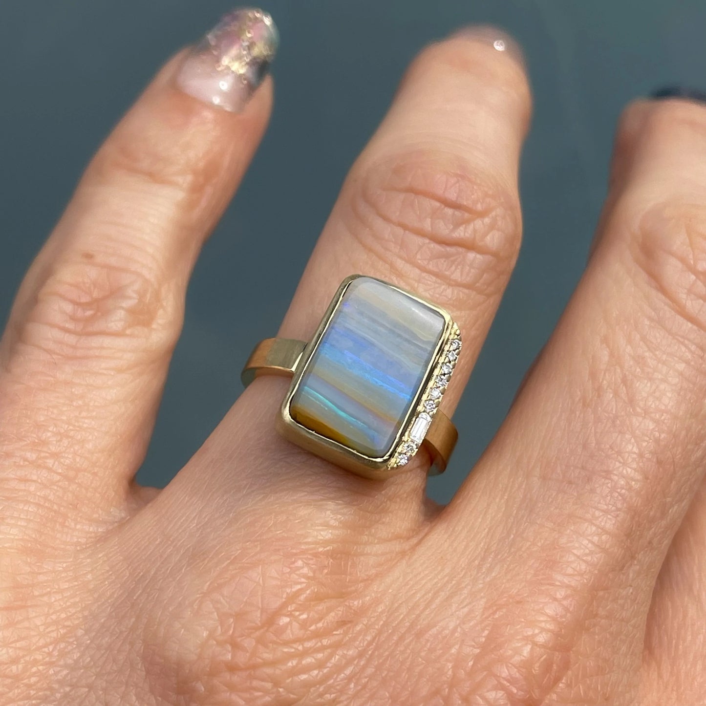 An Australian Opal Ring by NIXIN Jewelry with a blue opal and diamonds is modeled on a hand.