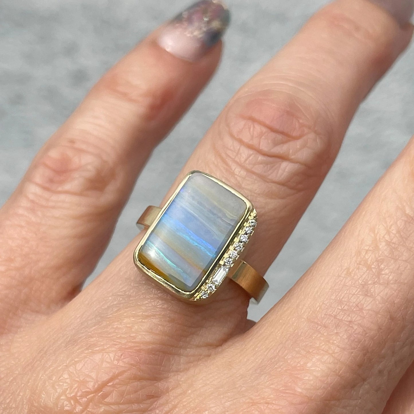 An Australian Opal Ring by NIXIN Jewelry modeled on a hand showing the blue opal and baguette diamond.