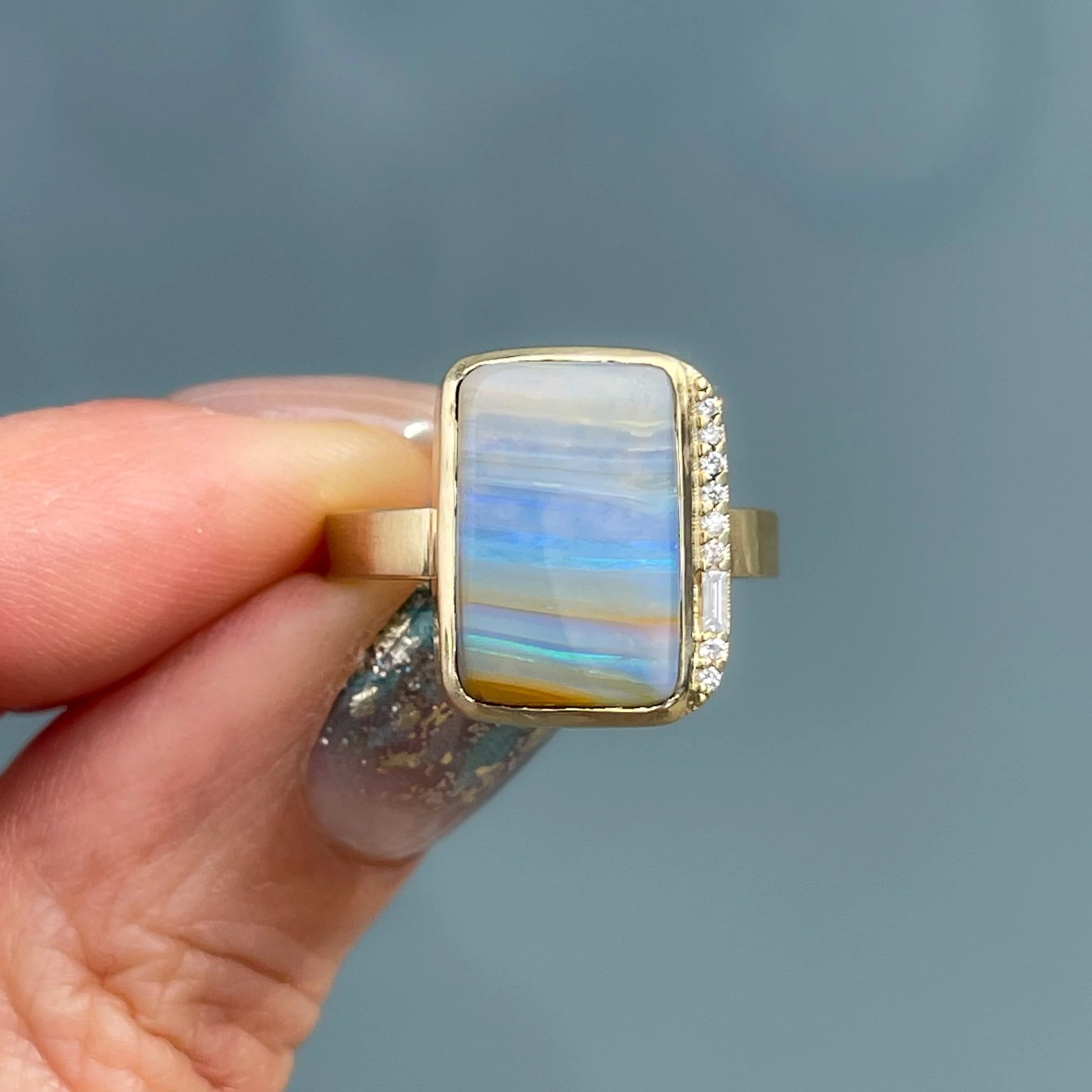 An Australian Opal Ring by NIXIN Jewelry with a Boulder Opal set in gold is held in front of a frosted glass backdrop.