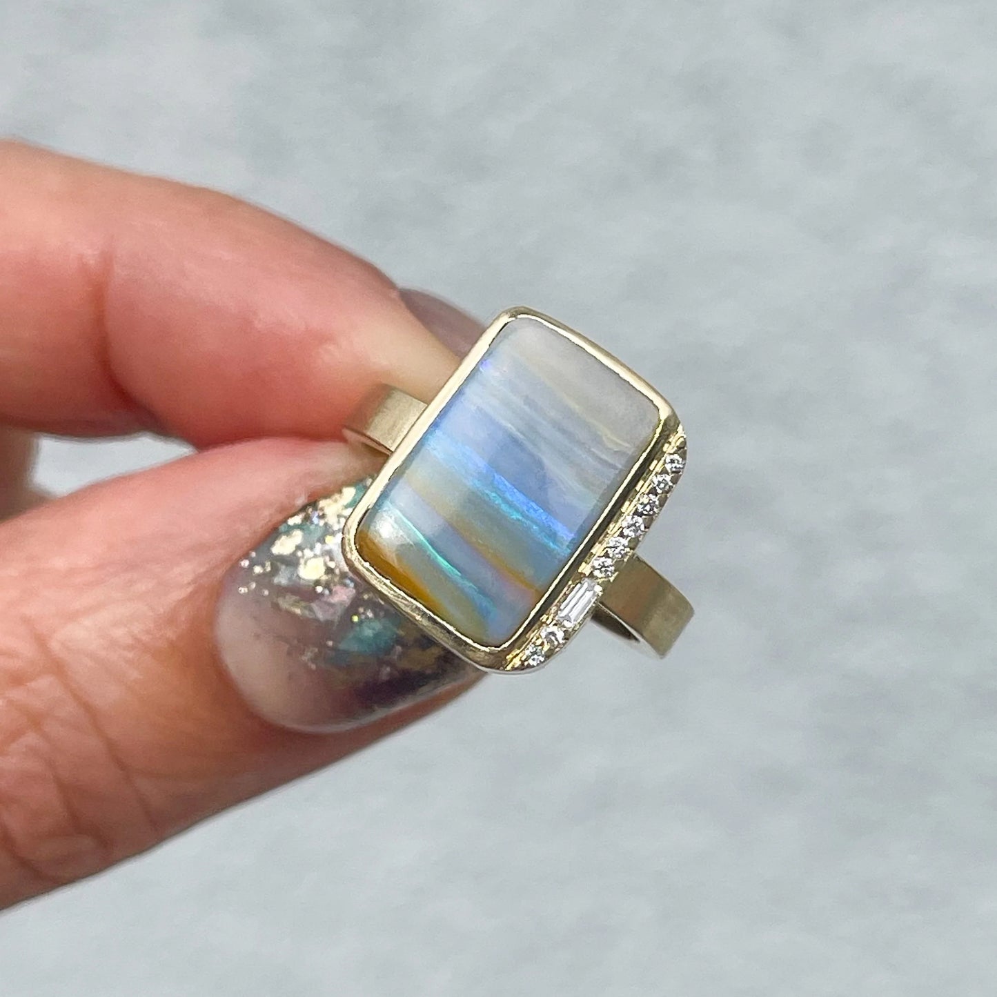 An Australian Opal Ring by NIXIN Jewelry with a blue green opal and pave diamonds is held in front of a grey wall.