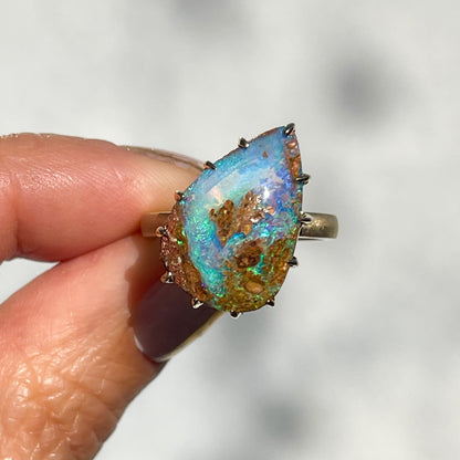 A NIXIN Jewelry Australian Opal Ring with a pear shaped opal set at an angle. The wood opal ring is held up in the sunlight.