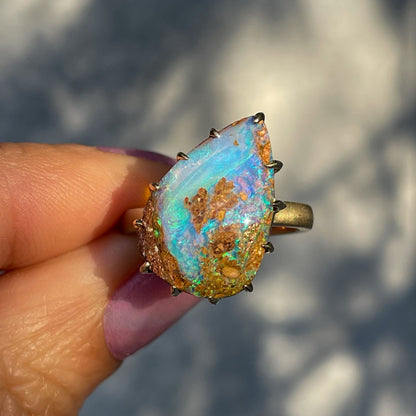 A NIXIN Jewelry Australian Opal Ring made in yellow gold with a wood fossil opal. The blue opal ring is held up in sunlight.