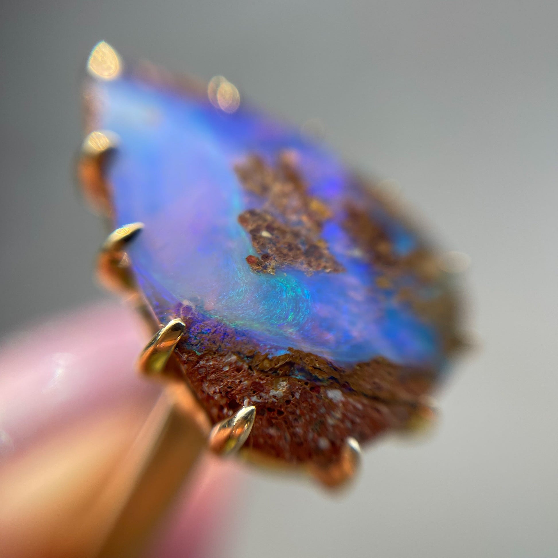 A magnified shot of a NIXIN Jewelry Australian Opal Ring in the shade, emphasizing its blue and purple tones. Shot at an angle featuring its 14k gold prong setting.