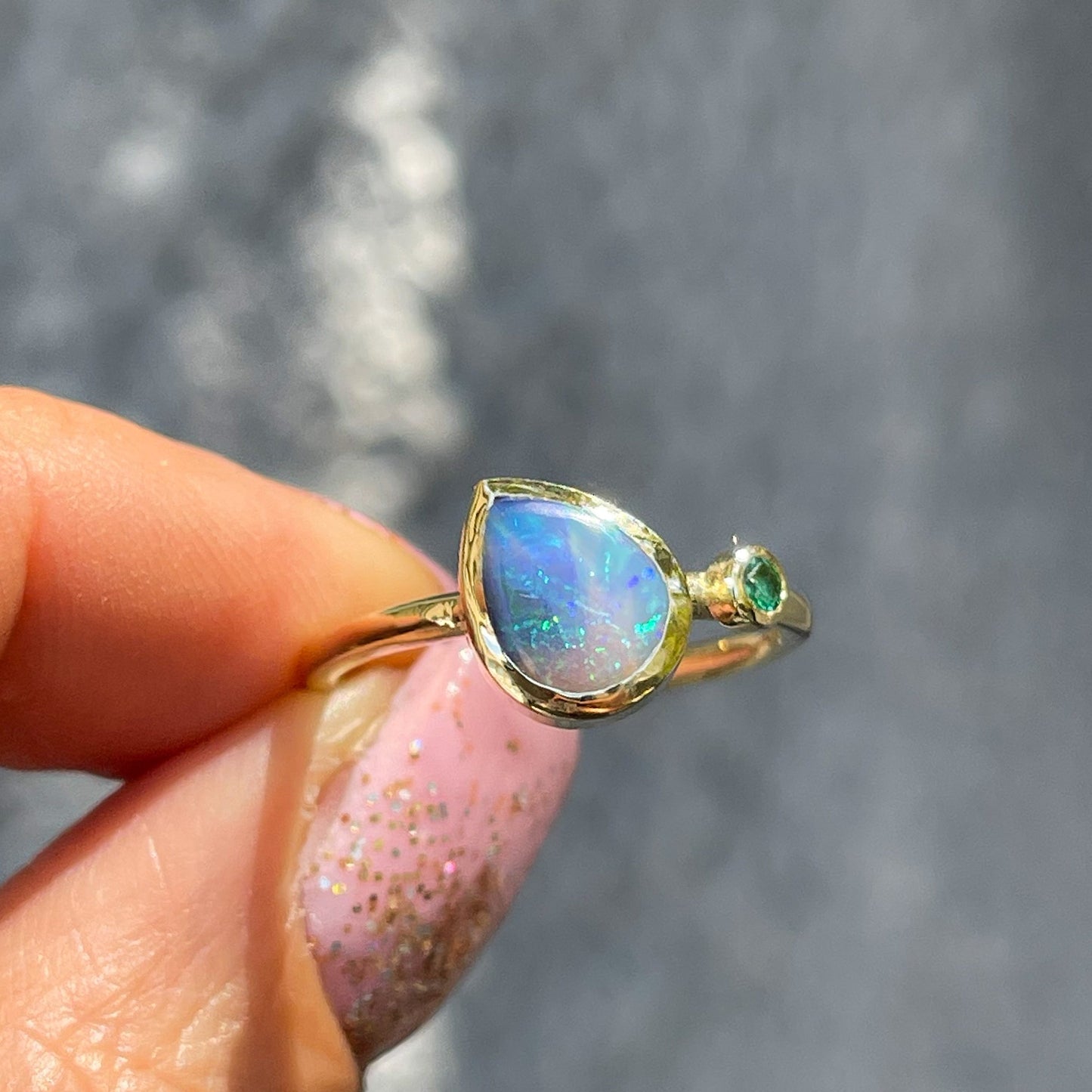 An Australian Opal Ring by NIXIN Jewelry held in front of a gray wall. A 14k gold emerald and opal ring.