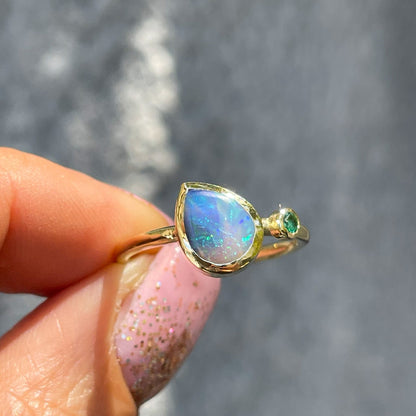 An Australian Opal Ring by NIXIN Jewelry held in front of a gray wall. A 14k gold emerald and opal ring.