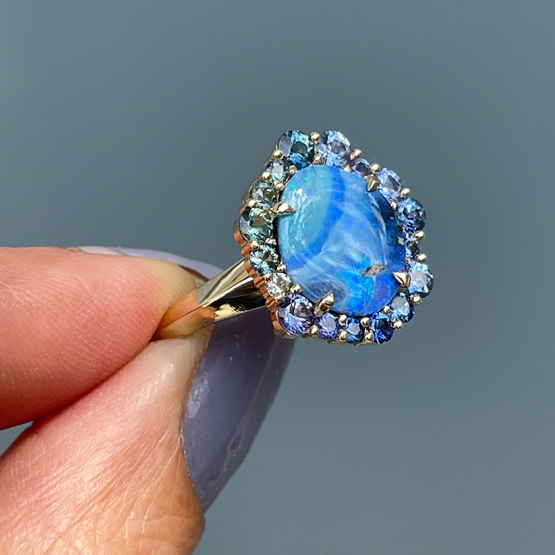 Australian Opal Ring by NIXIN Jewelry with an ombre halo of blue and green sapphires. Ring is held sideways in the shade.
