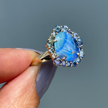 Australian Opal Ring by NIXIN Jewelry with an ombre halo of blue and green sapphires. Ring is held sideways in the shade.